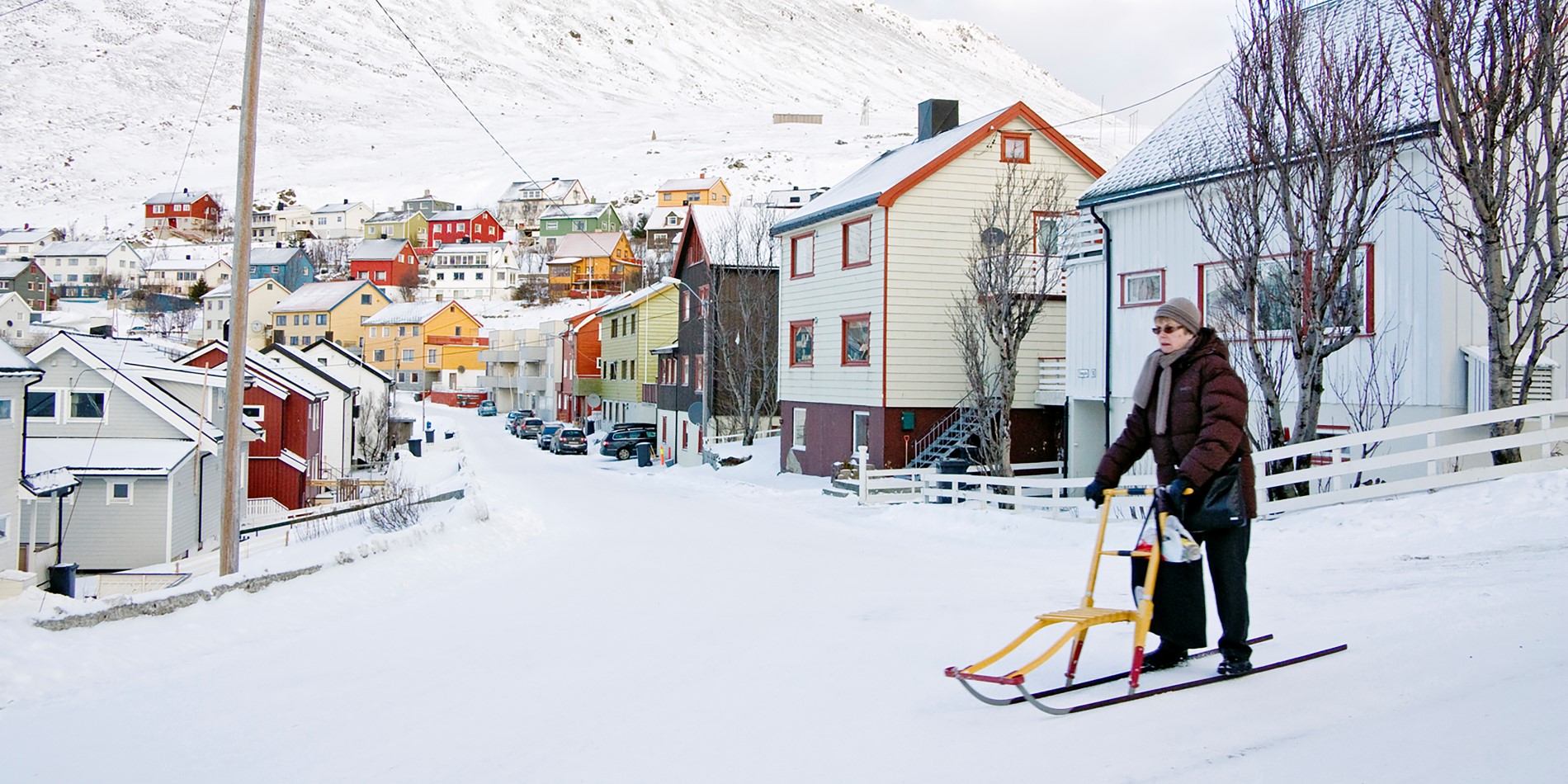Foto einer älteren Frau auf einer verschneiten Straße. Im Hintergrund sieht man schöne, bunt gestrichene kleine Holzhäuser.