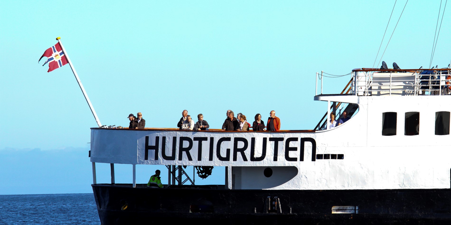 MS Nordstjernen, das schwarz-weiße Schiff, fährt mit im Wind wehender Flagge. Im hinteren Bereich des Schiffes stehen Passagiere, die die Aussicht genießen