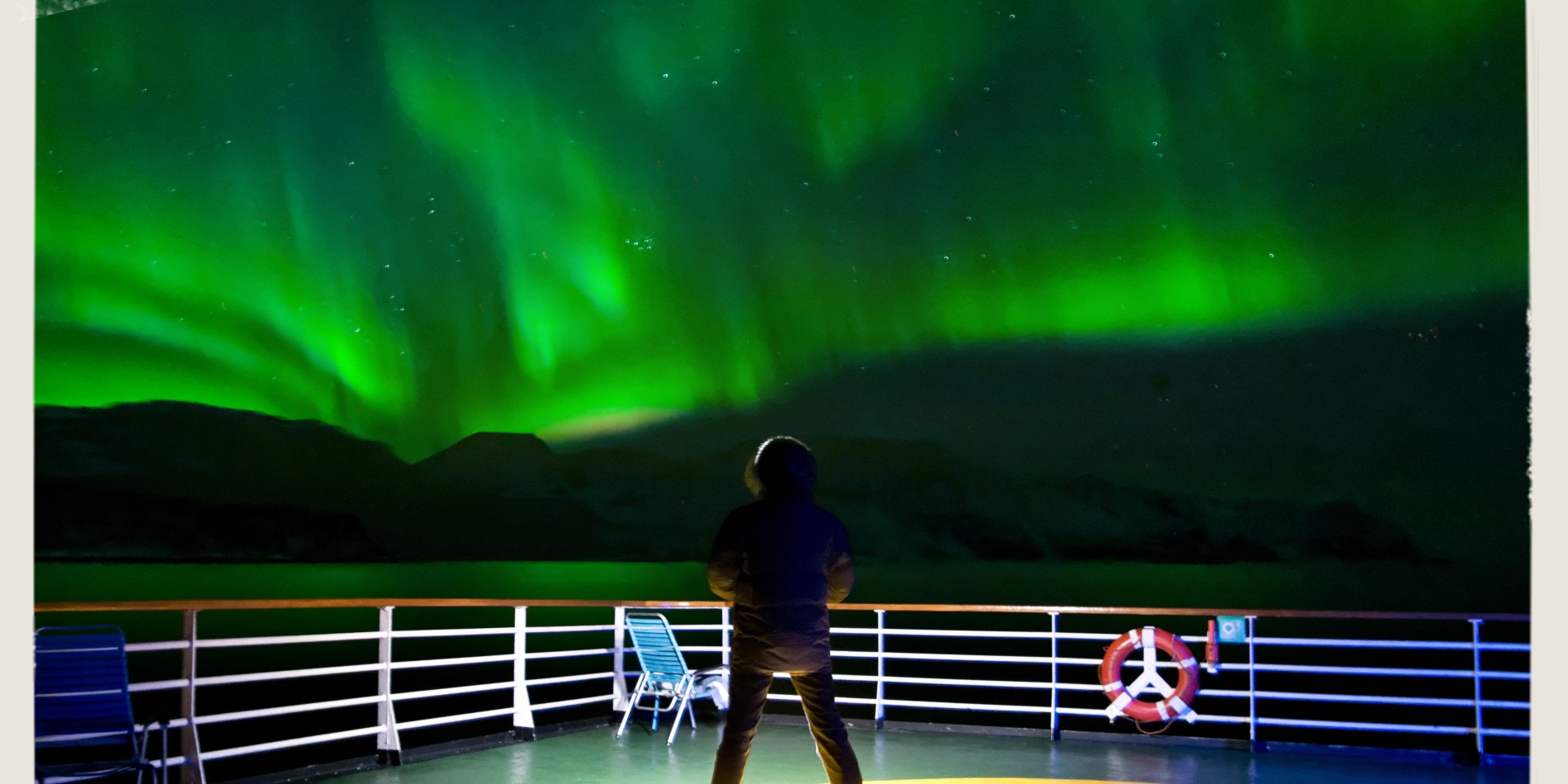 Mann an Deck eines Schiffs von Hurtigruten, das auf der klassischen Postschiffroute unterwegs ist. Über ihm schimmert das Nordlicht grünlich am Himmel.
