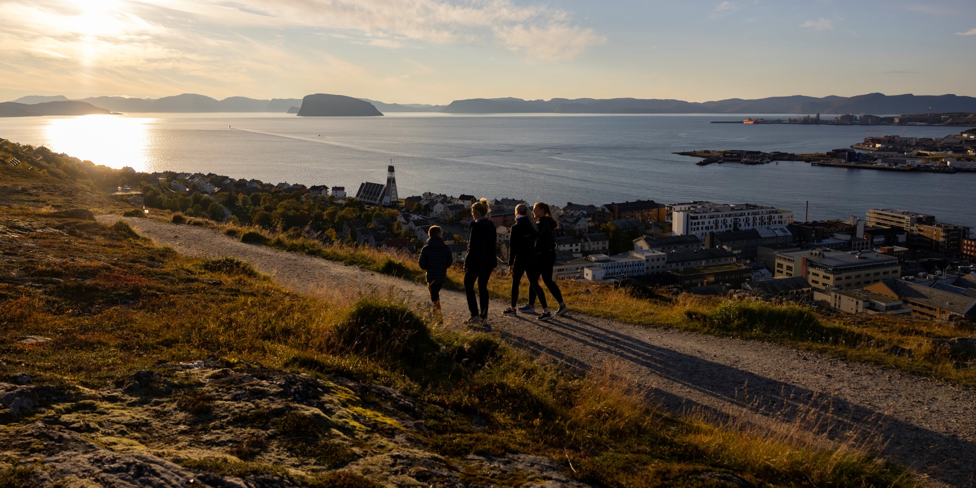 Vier Personen spazieren bei Sonnenuntergang durch eine Hügellandschaft. Unter ihnen liegt die Stadt Hammerfest